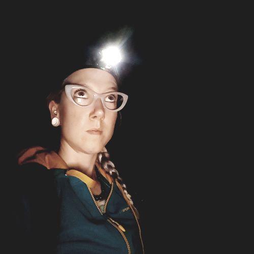 Woman wearing white cat eye glasses looks up. She is surrounded by darkness and has a lit headlamp on her head.
