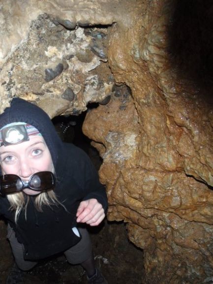 White woman wearing a black hoodie with the hood up emerges from an opening inside a cave. She is wear a head lamp and is carrying her sunglasses in her mouth.
