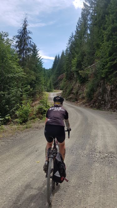 A rear view of a woman clad in black cycling gear riding down a winding gravel path surrounded by evergreen trees. A black pannier hangs off the right rear side of the bike.