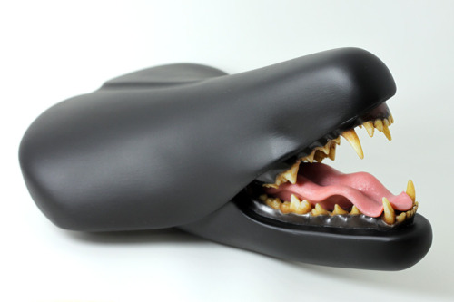 Black bicycle saddle with a jaw of a carnivore below the saddle nose. A wavy pink tongue rests between the lower rows of teeth.