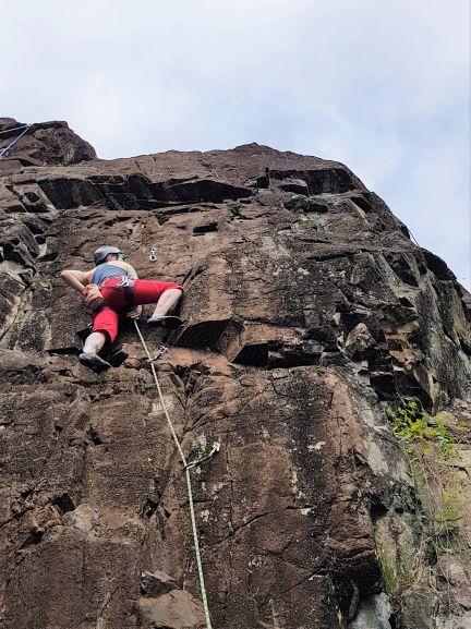 Laura stands hands-free on a rock face. A rope and two quickdraws are visible below her. She is wearing red capris, a blue tank top and a helmet. An orange chalk bag hangs off the back of her climbing harness.