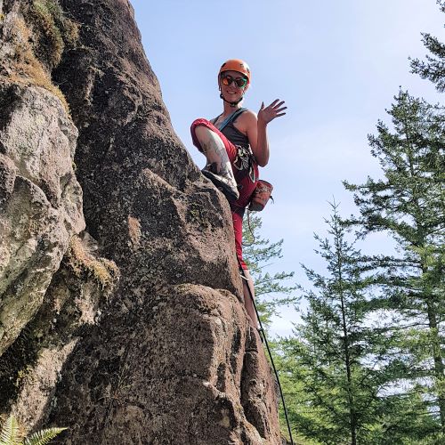 Laura waves from the middle of an eighty degree rock face. She is wearing an orange helmet, sunglasses, blue tank top, red capris and cllimbing shoes. An orange chalkbag hangs off the back of her harness. Trees are visible in the background.