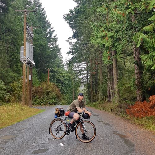 Ed poses on his bicycle on a wet road surrounded by trees. Ed is wearing a tiny cap, t-shirt, and shorts. Telephone poles can be seen on the left.