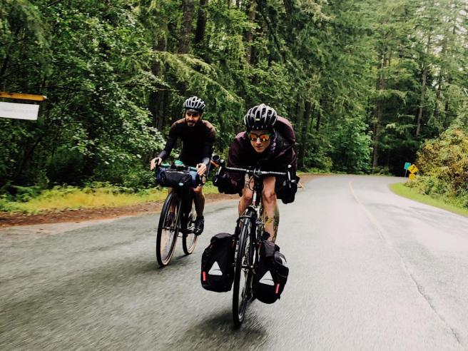 Laura gets aero on her bike while Yann follows behind. Laura is wearing a black helmet, sunglasses with reflective orange lenses and shorts. Her front panniers hang from the front rack. Yann, on the left, is dressed in all-black and is toting his gear in a handlebar bag. The road is wet and surrounded by trees.