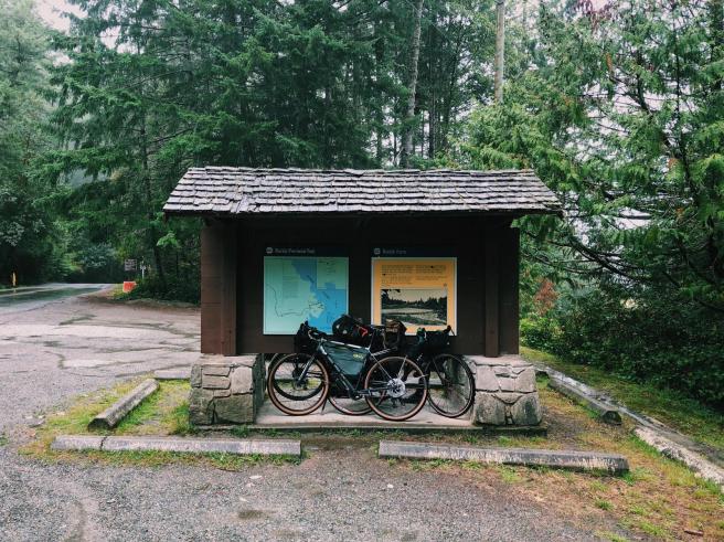 Two bicycles lean against a mini shelter. On the shelter, behind the bicycles is a map of Ruckle Provincial Park. Two concrete parking barriers lie in front of the shelter. Trees are visible in the background.