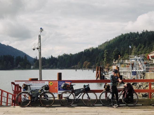 Three bikes lean against the railings of a dock. Laura stands on the right, in front of her bicycle, removing her arm warmer. The Fulford Harbour ferry terminal is visible in the background.