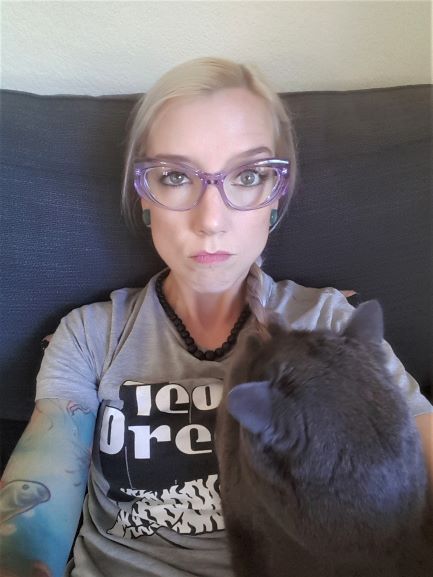 Laura stares directly at the camera while her fat, grey cat chews on her lavender-tinged braids, which are pulled to her left side. She is wearing translucent purple cat eye eyelgass frames, black necklace, and a heather grey t-shirt where the letters "Tea Dre" are visible (Team Dream).