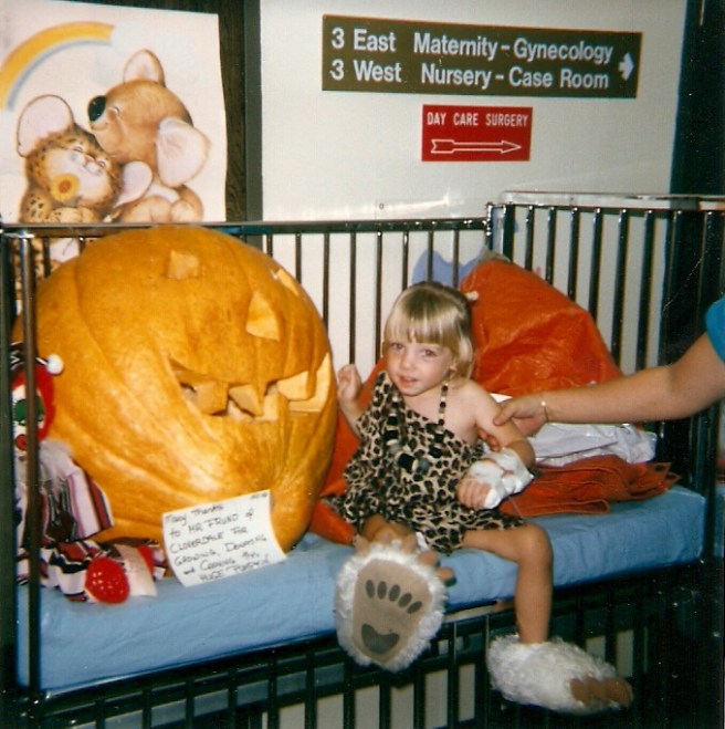A three year-old Laura in a leopard print Halloween costume and fuzzy white bear claw slippers is seated next to a giant jack-o-lantern. Her left arm appears to be wrapped in a half-cast, which is actually an IV port. The arm of an adult is grabbing her upper arm.