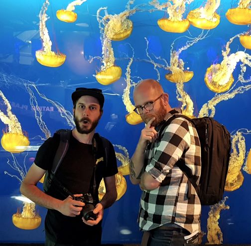 Yann and Ed stand before a tank full of Pacific sea nettles. Yann is dressed in black and his mouth is agape. In his hands is a DSLR camera. Ed is dressed in black and white flannel and is posing with his right finger under his chin.