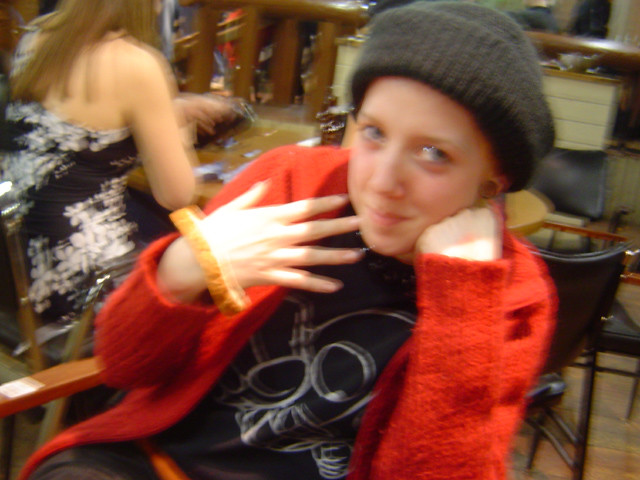 A blurry photo of Laura resting her head on one fist while showcasing a slice of bread around her other wrist. She is wearing a thick black toque, a red zippered sweater with a black t-shirt featuring Ultraman underneath.