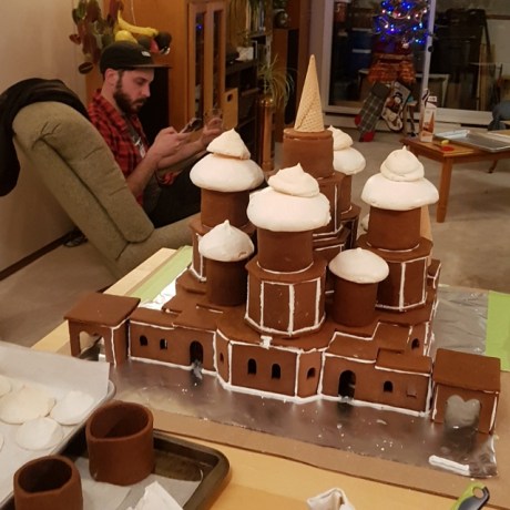 Gingerbread house pre-decoration. An inverted waffle cone can be seen on the top. To the left of the structure, is an out-of-focus Yann staring at his phone.