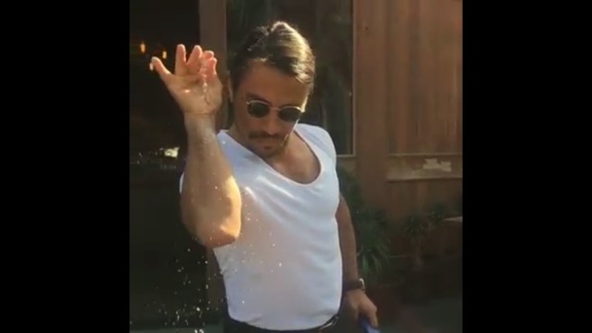 A man in dark sunglasses and wearing a white low-cut t-shirt sprinkles salt from his index finger and thumb pinched together. He is known as the meme 