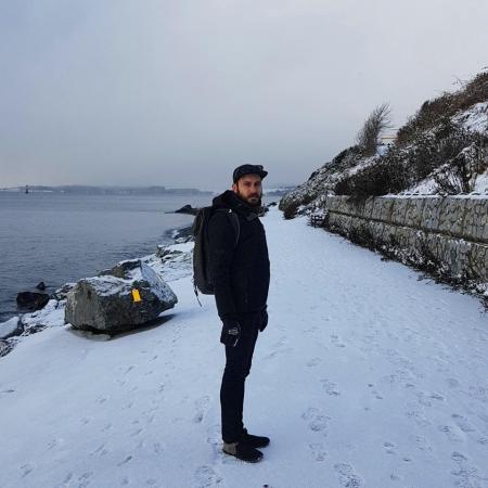 Yann, dressed in all-black and wearing a backpack, grimaces at the camera. The path by the beach is blanketed in snow.