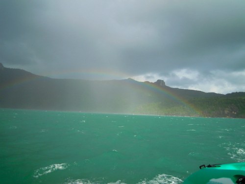 A low full arc of rainbow from the green-blue sea. Heavy clouds are visible above.