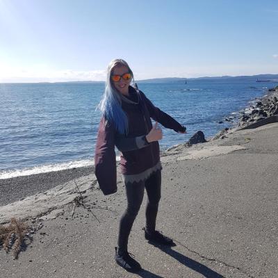 Laura smiles at the camera and gives a thumbs up through the pit vent of her purple jacket, giving the impression that she has three arms. The ocean is visible in the background.
