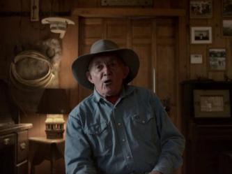 An old white man dressed in a denim blue button up and cowboy hat is mid-sentence.