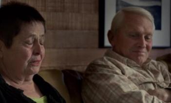 The woman on the left has short, dark hair. Her eyes are closed while she's mid-sentence. Next to her is her white haired husband with his arms crossed. He has a slight, contemplative grin on his face.