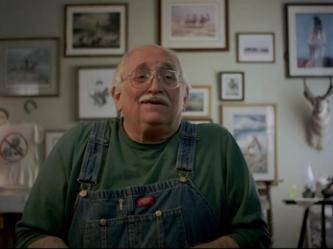 An obese bald old man with a white moustache and aviator glasses looks to the left. He is wearing a dark green t-shirt, blue coveralls. The wall behind him is covered in framed photos. On the right is the head of what appears to be an antelope. On the left is a white t-shirt, and above that, two horseshoes.