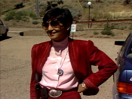 Sheela, a small woman with dark, short hair smiles with one hand on her hip. She's wearing oversized tinted glasses, burgandy jacket, pink blouse, a large silver buckle. Around her neck is a long, beaded pendant holding a portrait of Bhagwan.