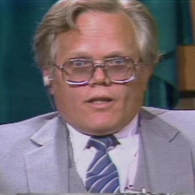 A broad-faced wide-eyed man with thick aviator glasses has his mouth slightly agape. He's wearing a light gray suit with a matching vest underneath.