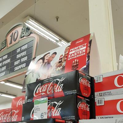 A display sign sits on top of boxes of Coca-Cola. The sign features a diverse group of friends holding up bottles of coke. The sign reads: Perfect with Easter Gatherings.