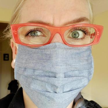 One eyebrow is slightly raised while I have a pained look in my eyes. I am wearing orange eyeglasses and a light blue mask.