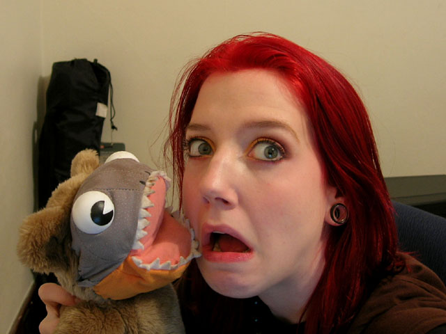 An old photo of me from when I had bright red hair. I am holding up the aforementioned piranha-faced bear plushy. My mouth is open.