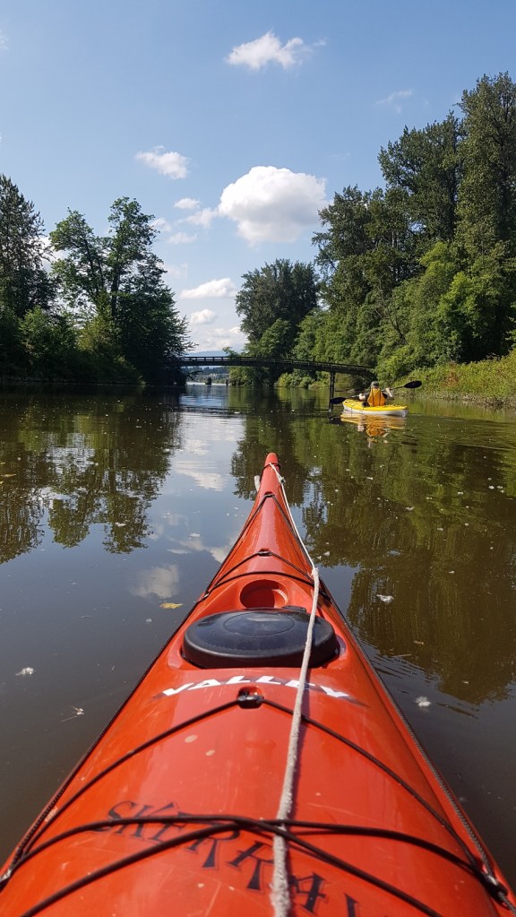 Yann's POV from a red kayak showing me paddling a yellow kayak in front, to the right. A bridge is visible in the background.