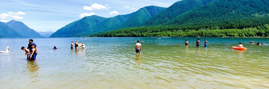 Yann in swim trunks stands knee-deep in the middle of a lake which is dotted with a few waders and even fewer swimmers. Tree-blanketed mountains are visible in the background.