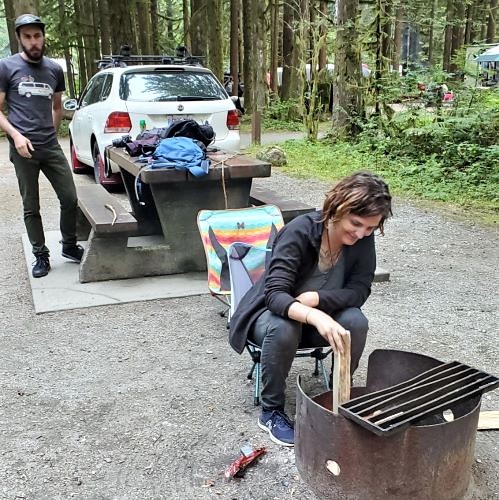 Zoee is seen in the foreground putting kindle in a fire pit. She has short brown hair and is wearing a black fleece jacket, black jeans, and navy blue New Balance sneakers. Yann stands to the left in the background scoping Zoee's fire starting technique. He is wearing a black hat, a dark grey t-shirt with a white van on it and dark pants.