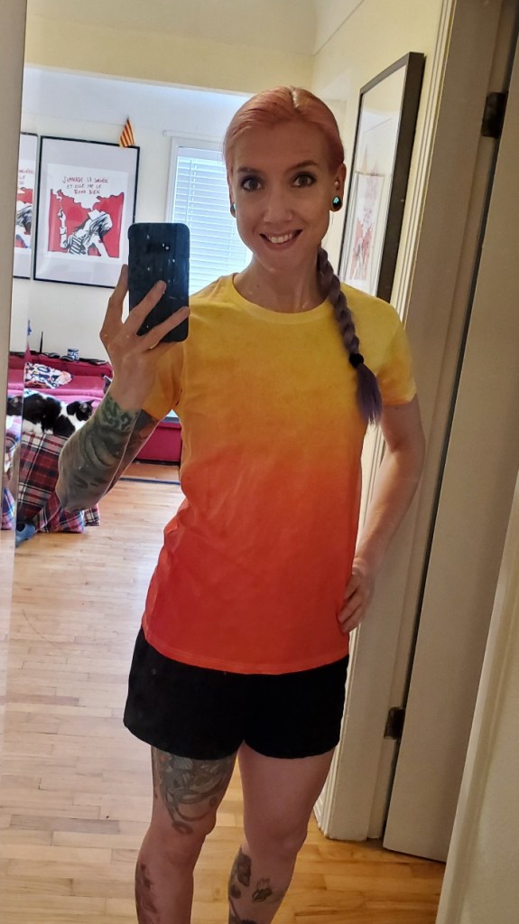 Laura bares her teeth in a smile. Her t-shirt is an orange to yellow ombre. Her hair is in a braid, pulled over one shoulder. A black and white cat can be seen sitting on the couch in the background.