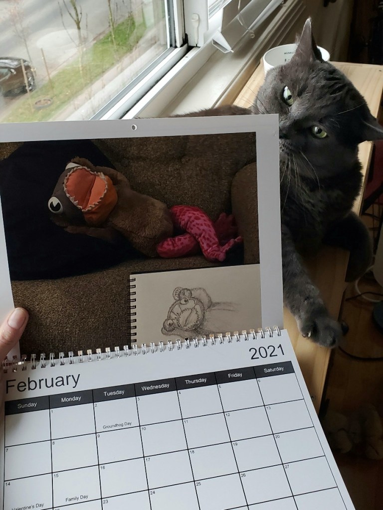 A calendar showing a plushie with the face of a piranha, body of a teddy bear, and legs of a pink frog lying on the couch with a sketchbook in front. The calendar is turned to the month of February. Behind the calendar is a curious grey cat.