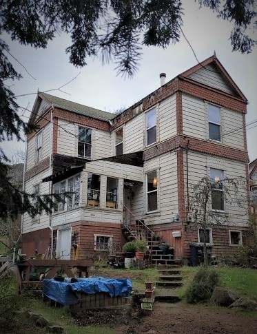 A three-storey wood panelled L-shaped building with wooden stairs curving from the main entrance to the dirt driveway. A blue tarp is visible in the lower left corner.