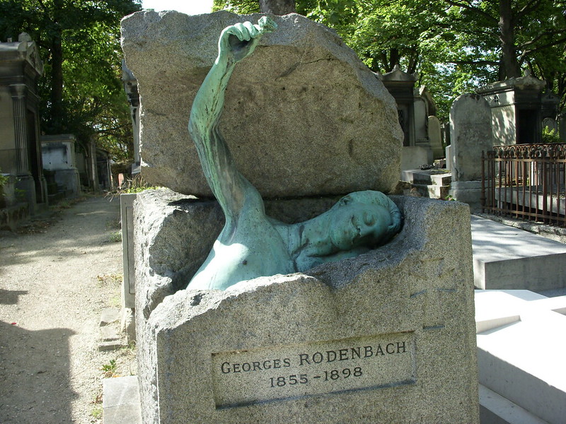 A patina-coated statue rises out of a granite block. Engraved on the headstone is: Georges Rodenbach 1855-1898.