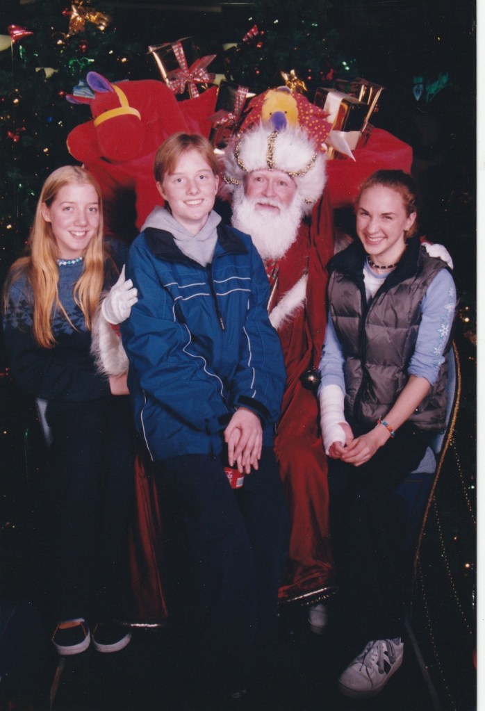 Three teenagers and a mall Santa. I'm on the left, dressed in dark blue. My hair is long, blonde and parted in the middle. Alana is int he middle, wearing a blue snowboarding jacket over a grey hoodie. She has short dark blonde hair. Erin is on the right wearing a puffy vest over a light blue baseball shirt. Her shirt sleeves are rolled up and reveals an arm in a plaster cast.
