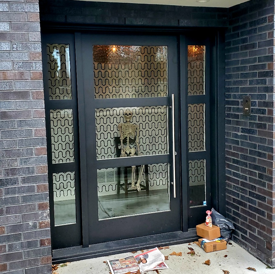 A skeleton sits on a wooden chair inside a house. Outside the door, a stack of parcels sit to the right of the door.