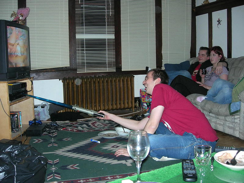 A lauging man in a burgandy t-shirt uses a collapsible light saber to reach the buttons on a VCR. On the TV monitor, a pornographic actor has her legs spread with one hand on her muff. Across from the tv is a heterosexual couple: the lady has a disturbed look on her face.