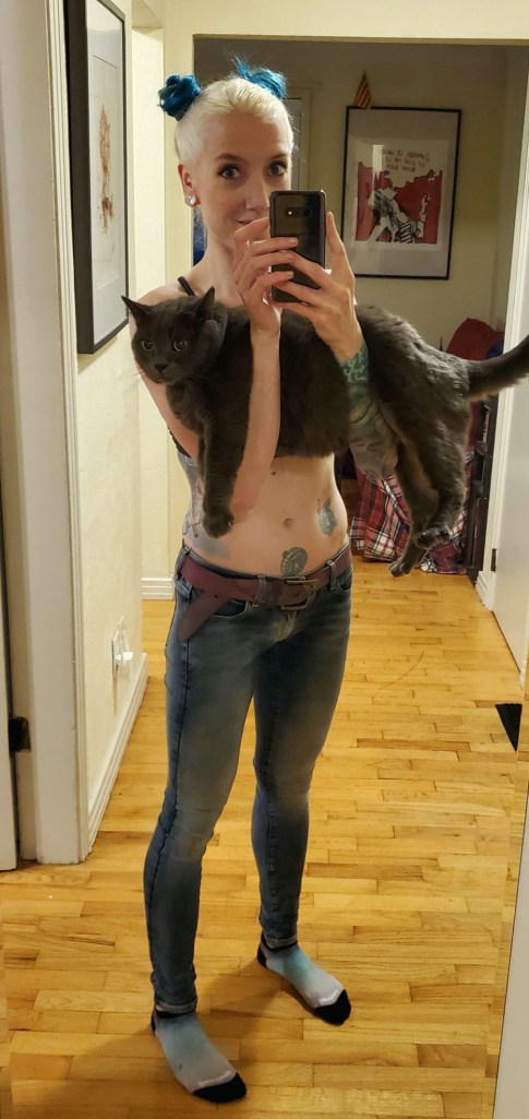 A mirror selfie. I am cradling a large grey cat while holding my phone with both hands. The cat is covering most of my torso. My bleach blonde hair is parted in the middle with two turquoise coloured buns on the side to resemble bear ears. I am wearing low rise blue demin and a lavender belt.