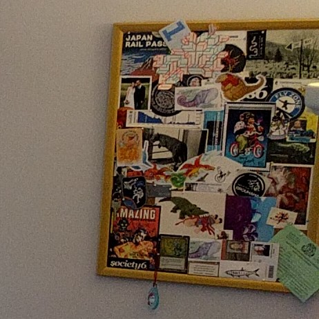 A grainy photo cropped from a larger photo. A gold framed corkboard is completely covered with miscellaneous artwork, postcards, stickers, and photos. The only text that is clear in the photo is: Japan Rail Pass at the top left, and AMAZING at the bottom left corner.