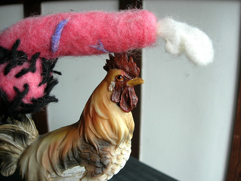 A pink needle felted cock resting on top of cock (rooster) figurine.