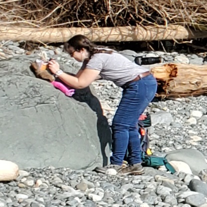 A close-up of Tammy bent over at the waist, trying to manipulate a plushie that's been placed on a boulder.